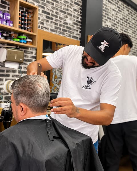 Barbeiro de boné finalizando o corte de um cliente grisalho com tesoura e pente