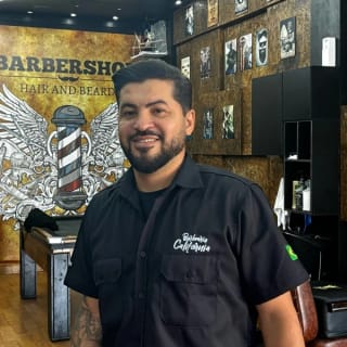 Maurício sorrindo, de barba, com camisa preta da Barbearia Califórnia, ao lado da mesa de sinuca