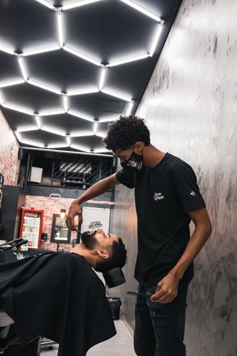 Barbeiro aparando a barba de um cliente na tesoura, sob o teto de luzes hexagonais